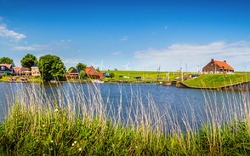 Panorama eines Flusses mit Ufergebäuden und grasbewachsenen Deichen.