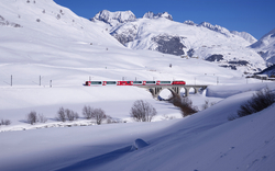 Ein roter Zug der Rhätischen Bahn, bekannt als Glacier Express, überquert ein Viadukt in einer verschneiten Berglandschaft auf der Albulalinie. Im Hintergrund sind schneebedeckte Berge und blauer Himmel zu sehen.