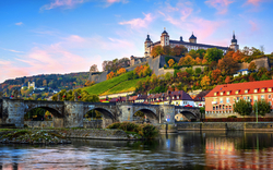 Festung auf einem Hügel mit Brücke im Vordergrund, herbstliche Bäume im Hintergrund.