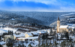 Winterliche Ansicht eines verschneiten Dorfes mit einer großen Kirche im Vordergrund, umgeben von schneebedeckten Häusern und einem Wald auf einem Hügel im Hintergrund. Der Himmel ist blau mit ein paar Wolken.