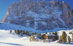 Hütte in einer verschneiten Berglandschaft vor einer großen Felswand unter blauem Himmel.