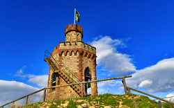 Ein kleiner, historischer Turm aus Stein mit einer Wendeltreppe außen, auf einem Hügel unter blauem Himmel mit Wolken, eine Flagge weht auf der Spitze.