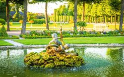 Springbrunnen mit Puttenfiguren in einem gepflegten Garten umgeben von Bäumen.