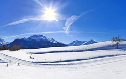 Panorama eines schneebedeckten Winterlandschaftsbildes mit einem strahlend blauen Himmel, einer leuchtenden Sonne, Bergen im Hintergrund und einer kleinen Holzhütte auf einem Hügel.