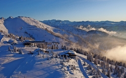 Verschneite Berglandschaft mit Skilift, Hütten und Bäumen im Sonnenaufgang, im Hintergrund ein Gebirgszug und ein blauer Himmel mit vereinzelten Wolken.