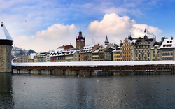 Panorama der schneebedeckten Kapellbrücke in Luzern, Schweiz, mit historischem Wasserturm, umliegenden Gebäuden und einem bewölkten Himmel im Hintergrund.