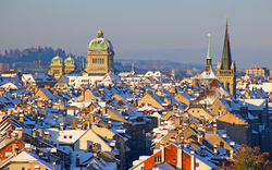 Verschneite Stadtansicht von Bern mit dem Bundeshaus und Kirchtürmen im Hintergrund, unter einem klaren blauen Himmel.