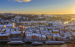 Verschneite Stadt an Flussufer bei Sonnenaufgang mit historischen Gebäuden.