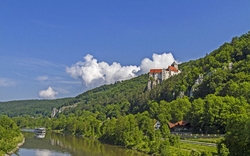 Ein Schloss auf einem bewaldeten Hügel über einem Fluss, an dem ein Boot fährt. Im Hintergrund ist ein blauer Himmel mit einigen Wolken zu sehen.
