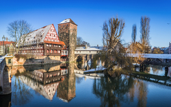 Panorama einer malerischen Flusslandschaft in der Altstadt von Nürnberg, Deutschland. Es zeigt Fachwerkhäuser und historische Gebäude, die sich im ruhigen Wasser des Flusses spiegeln. Eine Brücke verbindet die Ufer, und kahle Bäume sind am Flussrand zu sehen. Der Himmel ist klar und blau, was auf einen sonnigen Wintertag hindeutet.