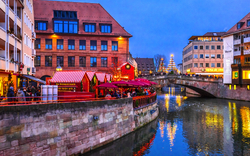 Nürnberger Christkindlesmarkt in der Abenddämmerung mit festlich beleuchteten Marktständen entlang eines Flusses, umgeben von historischen Gebäuden.