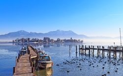 Ein See mit mehreren Booten an einem Steg, im Hintergrund schneebedeckte Berge und eine kleine Insel mit Gebäuden, viele Enten im Wasser.