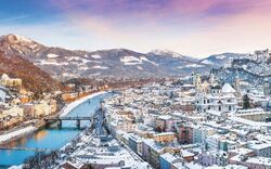 Panoramablick auf eine verschneite Stadt mit Fluss und Berglandschaft im Hintergrund bei Sonnenuntergang.
