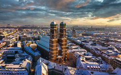 Winterliches Panorama von München mit der Frauenkirche im Schnee und Blick auf die Skyline bei Sonnenuntergang.