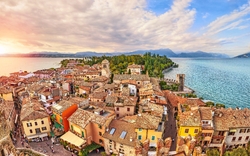 Panoramablick auf eine charmante italienische Stadt am Ufer eines großen Sees bei Sonnenuntergang, mit historischen Gebäuden, dicht aneinander liegenden Häusern und einem alten Schloss im Hintergrund.