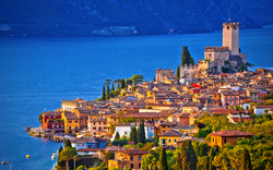 Blick auf eine malerische italienische Stadt am Seeufer mit einer mittelalterlichen Burg im Hintergrund. Die Stadt hat bunte Häuser und ist von üppigem Grün umgeben, während im Hintergrund ein großer See und bewaldete Berge zu sehen sind.