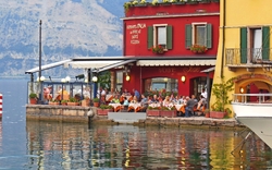 Eine malerische Szene eines italienischen Cafés oder Restaurants am Rande eines Sees mit mehreren Menschen, die an Tischen mit orangefarbenen Tischdecken sitzen und die Aussicht genießen. Im Hintergrund sind bunte Gebäude und eine bewaldete Berglandschaft zu sehen.