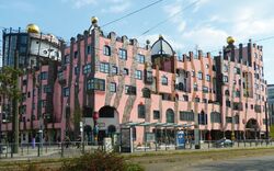 Ein ungewöhnliches, mehrstöckiges Gebäude mit rosa Fassade und unregelmäßigen Formen, bekannt als die Grüne Zitadelle in Magdeburg, entworfen vom Künstler Friedensreich Hundertwasser. Das Gebäude hat goldene Kuppeln auf dem Dach und eine Vielzahl von Fenstern in unterschiedlichen Formen.