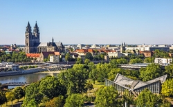 Panorama der Stadt mit einem großen gotischen Dom, Gebäuden am Flussufer und einer grünen Landschaft im Vordergrund.