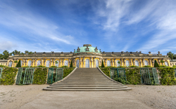Schloss Sanssouci in Potsdam, Brandenburg, ein historisches Schloss unter blauem Himmel, von grünen Büschen umgeben mit breiter Treppe im Vordergrund.