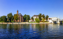Ein Fluss mit Bäumen und Gebäuden am Ufer, darunter eine Kirche und mehrere moderne Bauten, unter einem klaren blauen Himmel.