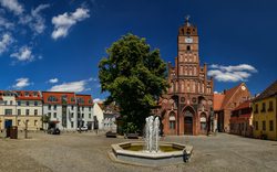 Ein historischer Marktplatz mit traditionellen Gebäuden, einer alten Kirche mit einem Turm und einer analogen Uhr. Vor der Kirche befindet sich ein Brunnen mit sprudelndem Wasser. Der Himmel ist blau mit einigen Wolken.