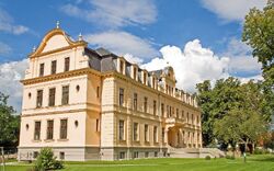 Ein historisches, cremefarbenes Gebäude mit mehreren Fenstern und einem aufwendig gestalteten Dach vor einem sonnigen Himmel mit Wolken, umgeben von gepflegtem Rasen und Bäumen.