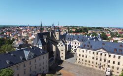 Luftaufnahme einer europäischen Stadt mit einem mittelalterlichen Schloss in der Mitte. Die Gebäude haben traditionelle Architektur mit Ziegeldächern. Der Himmel ist klar und blau am Tag.