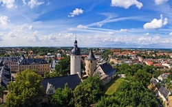 Panoramaansicht einer historischen Stadt mit einer Burg oder einem Schloss in der Mitte, umgeben von Bäumen und einer Vielzahl von Gebäuden, unter einem blauen Himmel mit weißen Wolken.