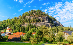 Eine malerische Ansicht eines kleinen Dorfes mit roten Dächern im Vordergrund und einem großen bewaldeten Sandsteinfelsen im Hintergrund unter einem strahlend blauen Himmel mit wenigen Wolken.