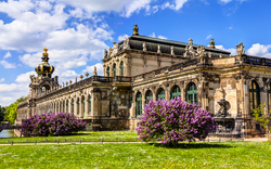 Bild des Zwingers in Dresden, einem barocken Museumskomplex und bedeutenden Wahrzeichen in Sachsen, Deutschland. Zu sehen sind das Kronentor, die langen Galerien und die Festungsmauer, umgeben von einem gepflegten Park mit Blumen und Brunnen.