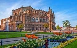 Ein großes, historisches Gebäude mit reicher Architektur, umgeben von bunten Blumenbeeten und einem klaren, blauen Himmel im Hintergrund.