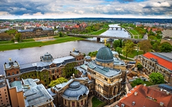 Das Bild zeigt eine Panoramaaufnahme von Dresden. In der Mitte des Bildes fließt die Elbe, über die eine Brücke führt. Im Vordergrund befinden sich historische Gebäude mit aufwendiger Architektur und Kuppeln. Der Himmel ist bewölkt und die Umgebung ist grün und städtisch geprägt.