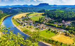 Luftaufnahme einer malerischen Landschaft mit einem Fluss, der sich durch grüne Wälder und Felder schlängelt. Im Vordergrund sind eine Stadt und landwirtschaftliche Flächen zu sehen. Im Hintergrund erstrecken sich bewaldete Hügel und ein klarer, blauer Himmel mit wenigen Wolken.
