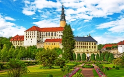 Ein großes, historisches Schloss mit rotem Dach und Turm, umgeben von gepflegten Gärten und blühenden Blumenbeeten unter blauem Himmel.