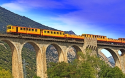 Gelber Zug überquert ein hohes Steinviadukt in einer bergigen Landschaft.