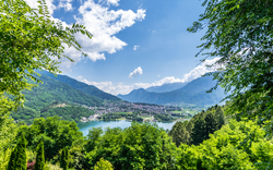 Landschaft mit Bergen, See und Bäumen unter blauem Himmel.