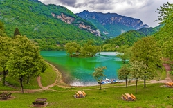 Grüner See inmitten von Bergen, umgeben von Bäumen, mit Booten am Ufer.