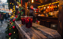 Zwei Gläser Glühwein auf einer Holzarbeitsplatte auf einem festlichen Weihnachtsmarkt mit Dekoration im Hintergrund.