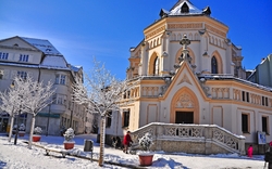 Winter in Rosenheim mit einer verschneiten Stadtlandschaft, in der ein historisches Gebäude steht. Bäume sind mit Schnee bedeckt und der Himmel ist klar und blau. Menschen bewegen sich im Vordergrund.