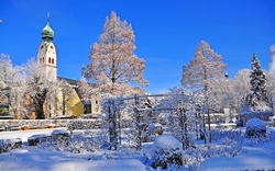 Eine verschneite Winterlandschaft in Rosenheim mit einem Kirchturm und schneebedeckten Bäumen im Vordergrund. Der Himmel ist klar und blau.