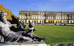 Eine beeindruckende Statue im Vordergrund eines großen, gepflegten Gartens mit einem historischen Schloss im Hintergrund. Die Szene wird von herbstlichen Bäumen umrahmt.
