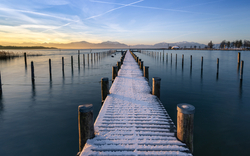 Schneebedeckter Steg am Chiemsee im Wintermorgenlicht, umgeben von ruhigem Wasser und Hintergrund der Alpen.