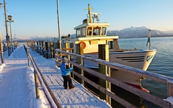 Ein Kind in winterlicher Kleidung geht auf einem verschneiten Steg auf ein Boot im Hafen zu, im Hintergrund sind schneebedeckte Berge und ein blauer Himmel zu sehen.