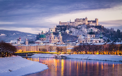 Winterliche Stadtansicht von Salzburg mit der Festung Hohensalzburg im Hintergrund, umgeben von schneebedeckten Gebäuden und einem Fluss im Vordergrund.