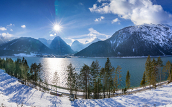 Verschneite Berglandschaft mit einem See im Tal, umgeben von schneebedeckten Bäumen, unter einem klaren blauen Himmel mit Sonnenschein.