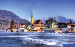 Dorfansicht am Ufer eines teilweise gefrorenen Sees mit Bergen im Hintergrund und einer Kirche mit hohem Turm in der Mitte der Szene.