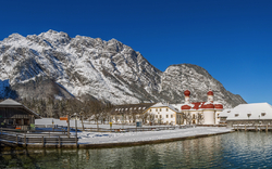 Panoramablick auf die verschneite Kirche St. Bartholomä auf der Halbinsel Hirschau am Königssee, umgeben von den winterlichen Bergen im Nationalpark Berchtesgaden in Bayern, Deutschland.
