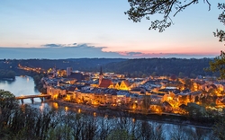 Abendliche Stadtlandschaft mit Fluss im Vordergrund und bewaldeten Hügeln im Hintergrund, Gebäude sind beleuchtet und der Himmel zeigt ein Abendrot.