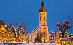 Eine verschneite winterliche Szene in einem bayerisch anmutenden Dorf. Im Vordergrund steht eine Kirche mit einer markanten Zwiebelkuppel, umgeben von beleuchteten Bäumen und Häusern. Die Szenerie ist in warmes Licht getaucht, kontrastierend zum blauen Abendhimmel.